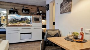 Eine moderne Küche mit weißen Schränken, einer Mikrowelle und einer Kaffeemaschine. Ein Holztisch mit einem Stuhl, einer Weinflasche und einer kleinen Pflanze sind ebenfalls vorhanden.