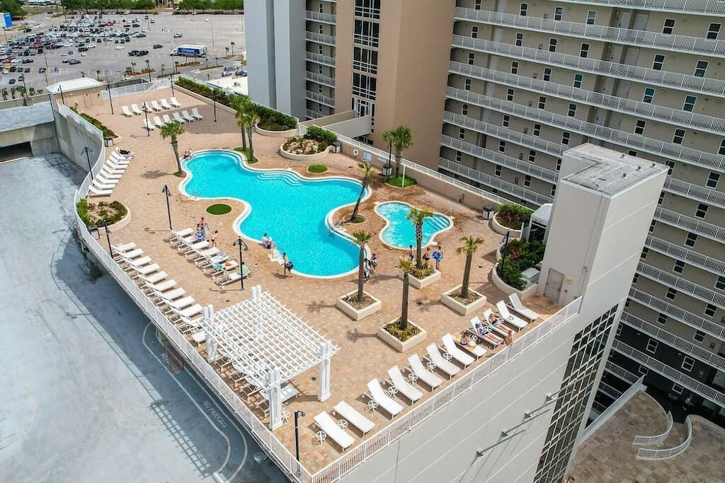 A rooftop pool area with multiple swimming pools, lounge chairs, and palm trees.