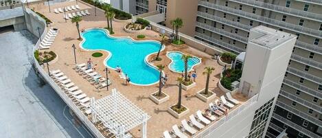 A rooftop pool area with multiple swimming pools, lounge chairs, and palm trees.
