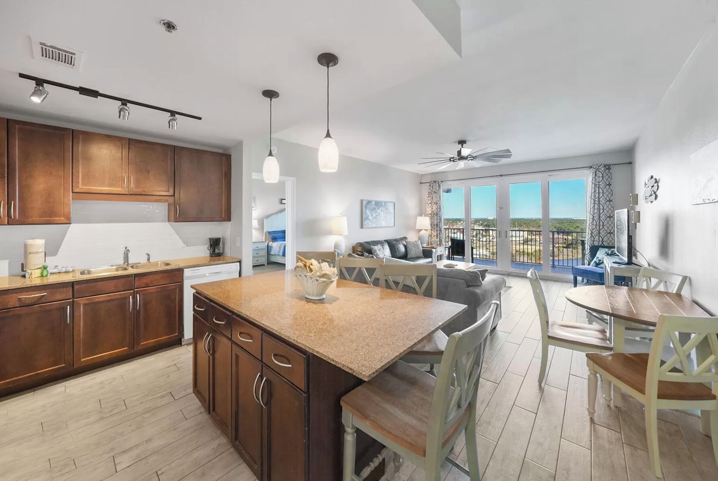 A modern kitchen with granite countertops, wooden cabinets, and pendant lighting. There is a dining area with a wooden table and chairs, and a large window offering a view of the outdoors.