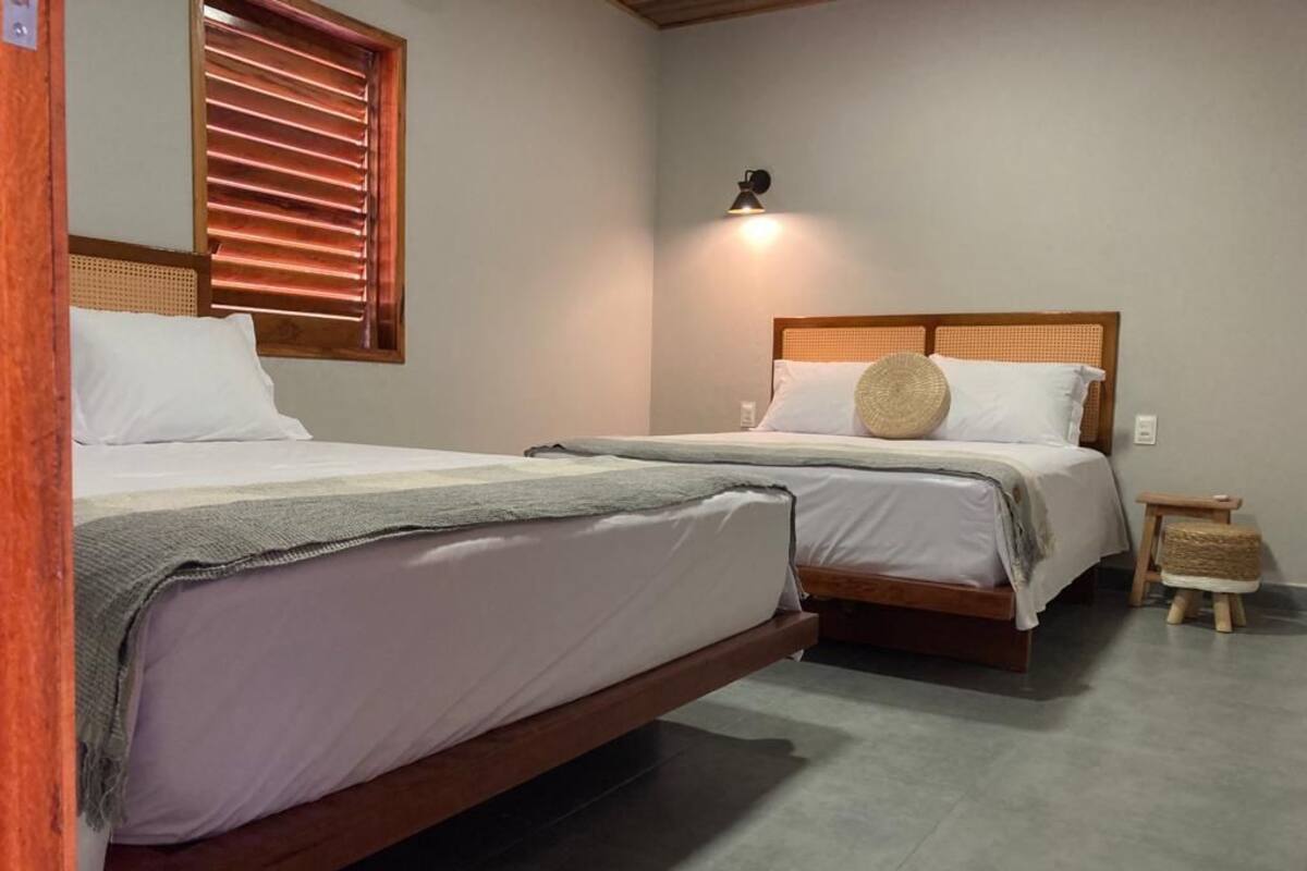 A bedroom with two beds, a small bedside table, a lamp, and a window with blinds.