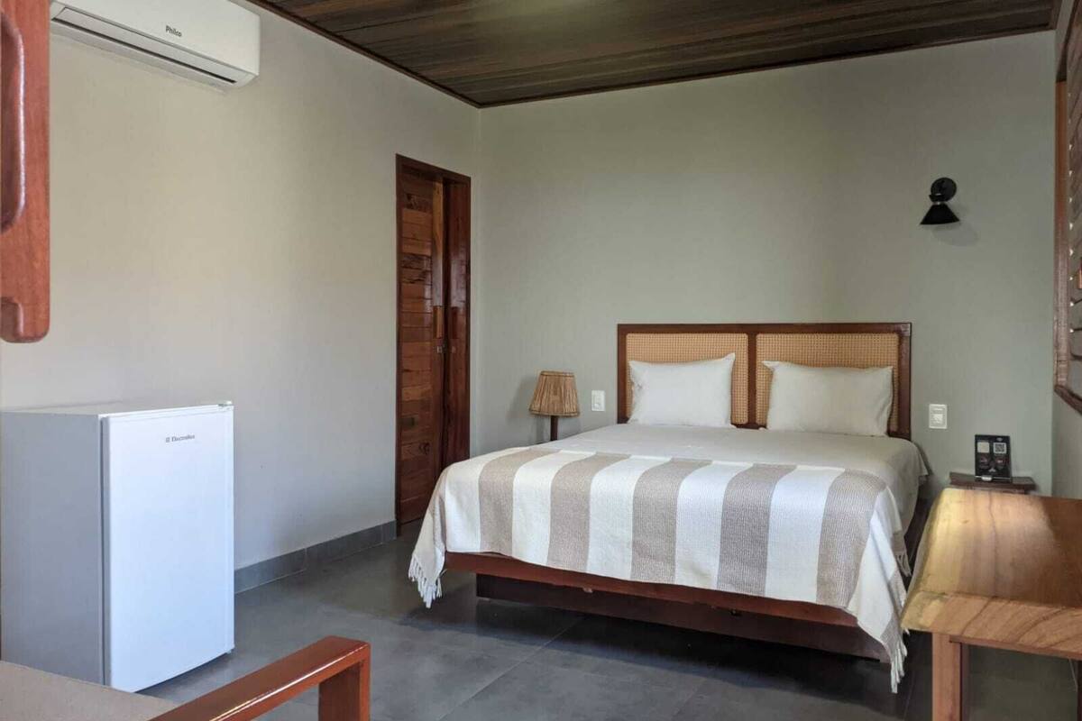 A bedroom with a bed, a bedside table, a lamp, a refrigerator, and a wooden door.