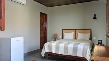 Quarto com cama, mesa de cabeceira, luminária, geladeira e porta de madeira.