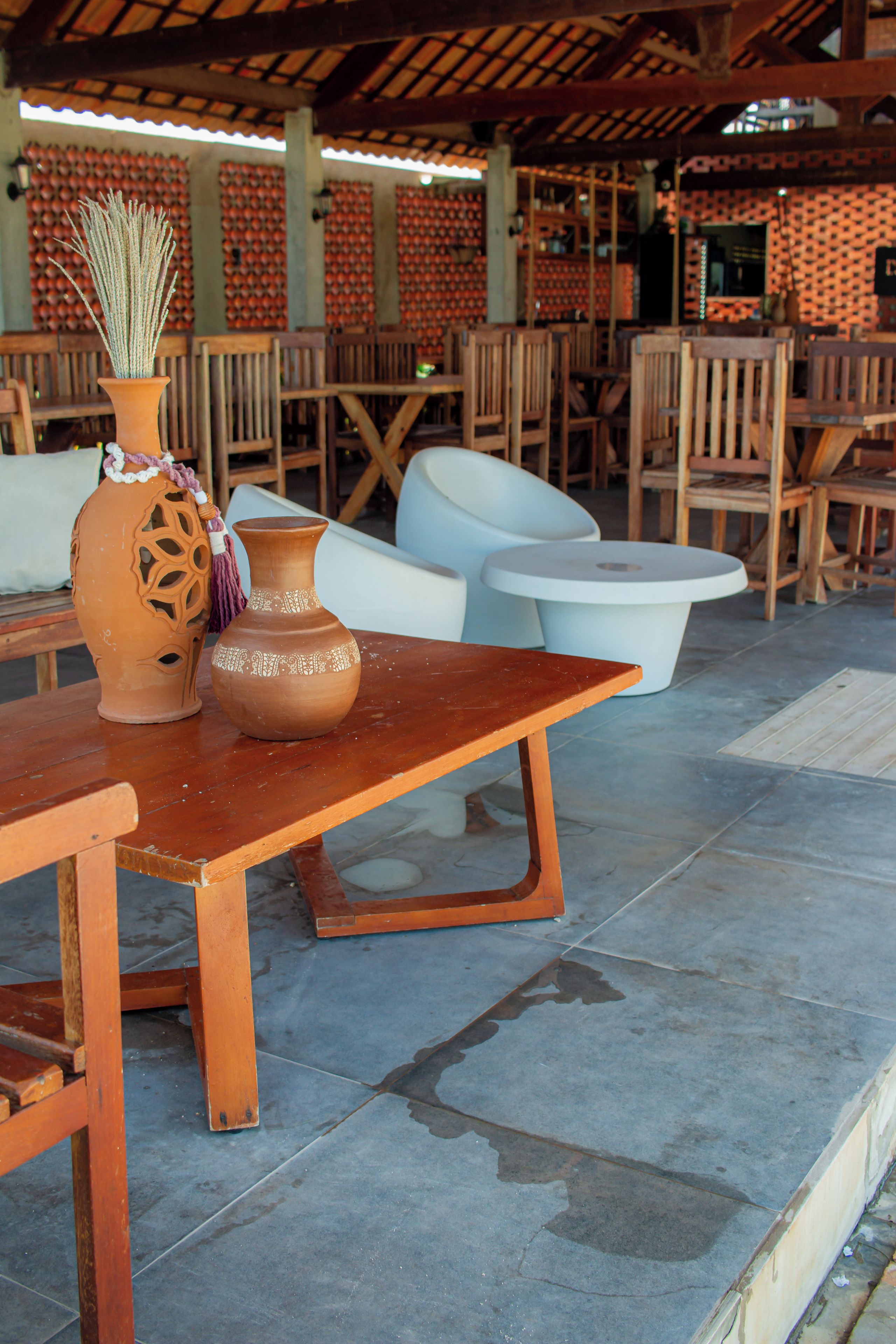 An outdoor seating area with wooden tables and chairs, a terracotta vase with a plant, and a patterned wall in the background.