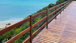 Una pasarela de madera con barandilla metálica que conduce a una playa de aguas azules y una orilla lejana.