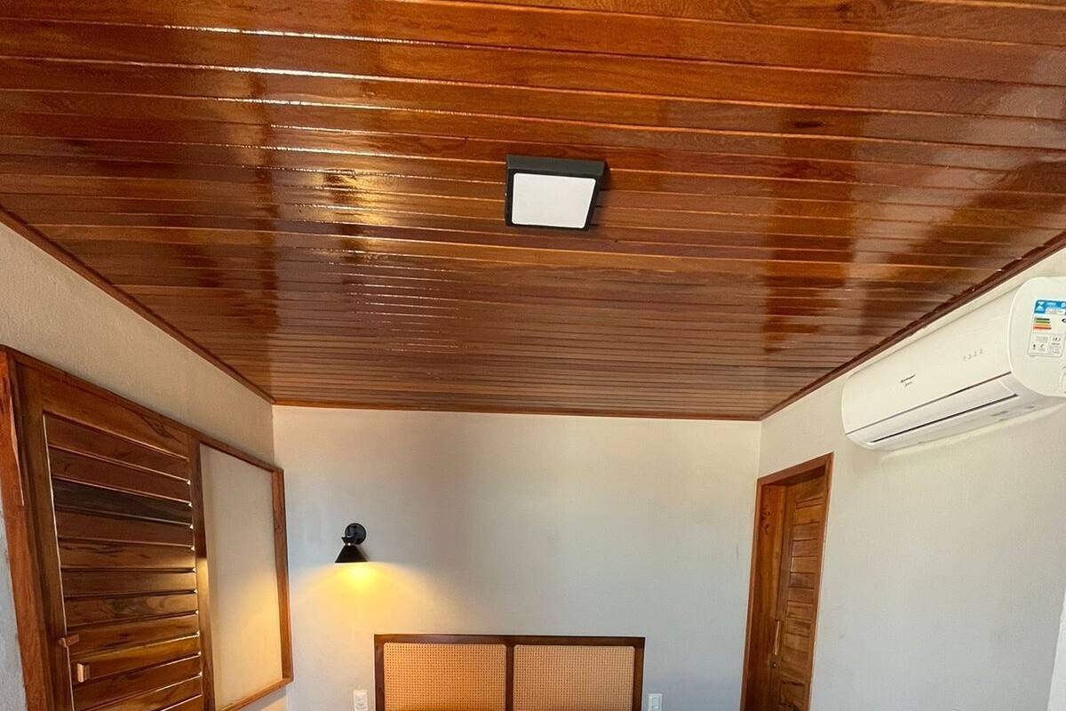 A hotel room with a wooden ceiling, a bed with a checkered bedspread, two pillows, a bedside table with a lamp, and an air conditioning unit on the wall.