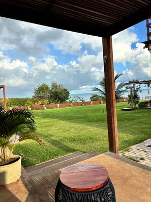 A covered outdoor area with a view of a grassy field and a wooden pergola. - Lyttos Bungalows (Icapuí)
