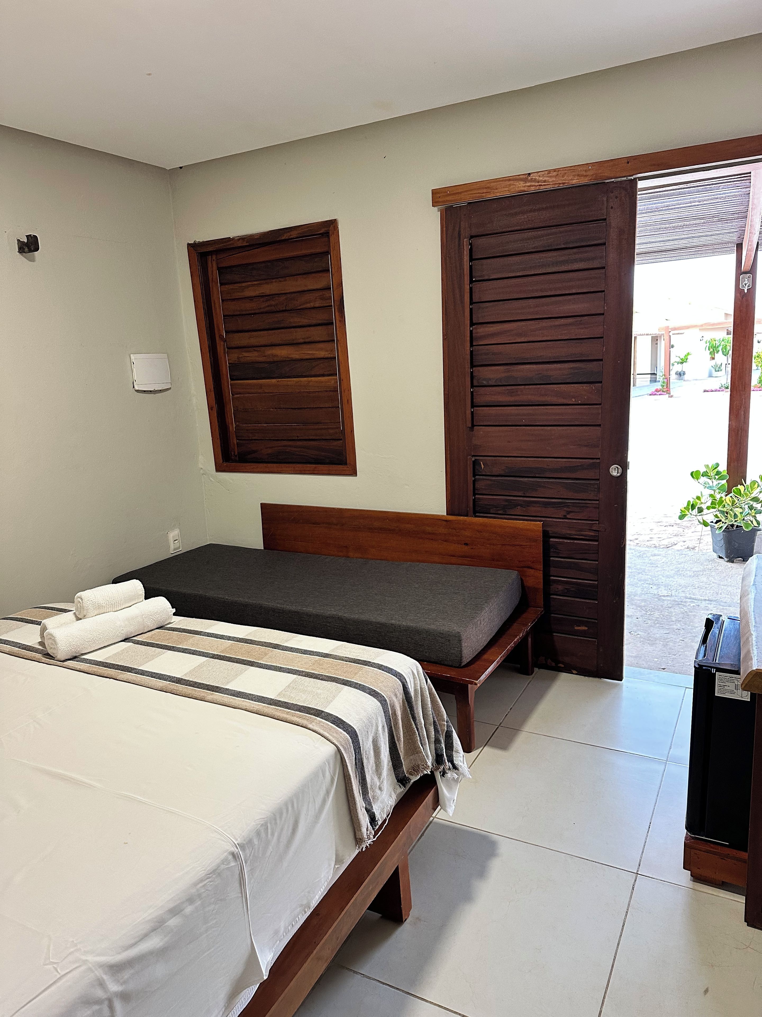 A bedroom with a bed, a bench, a wooden door, and a window with shutters.
