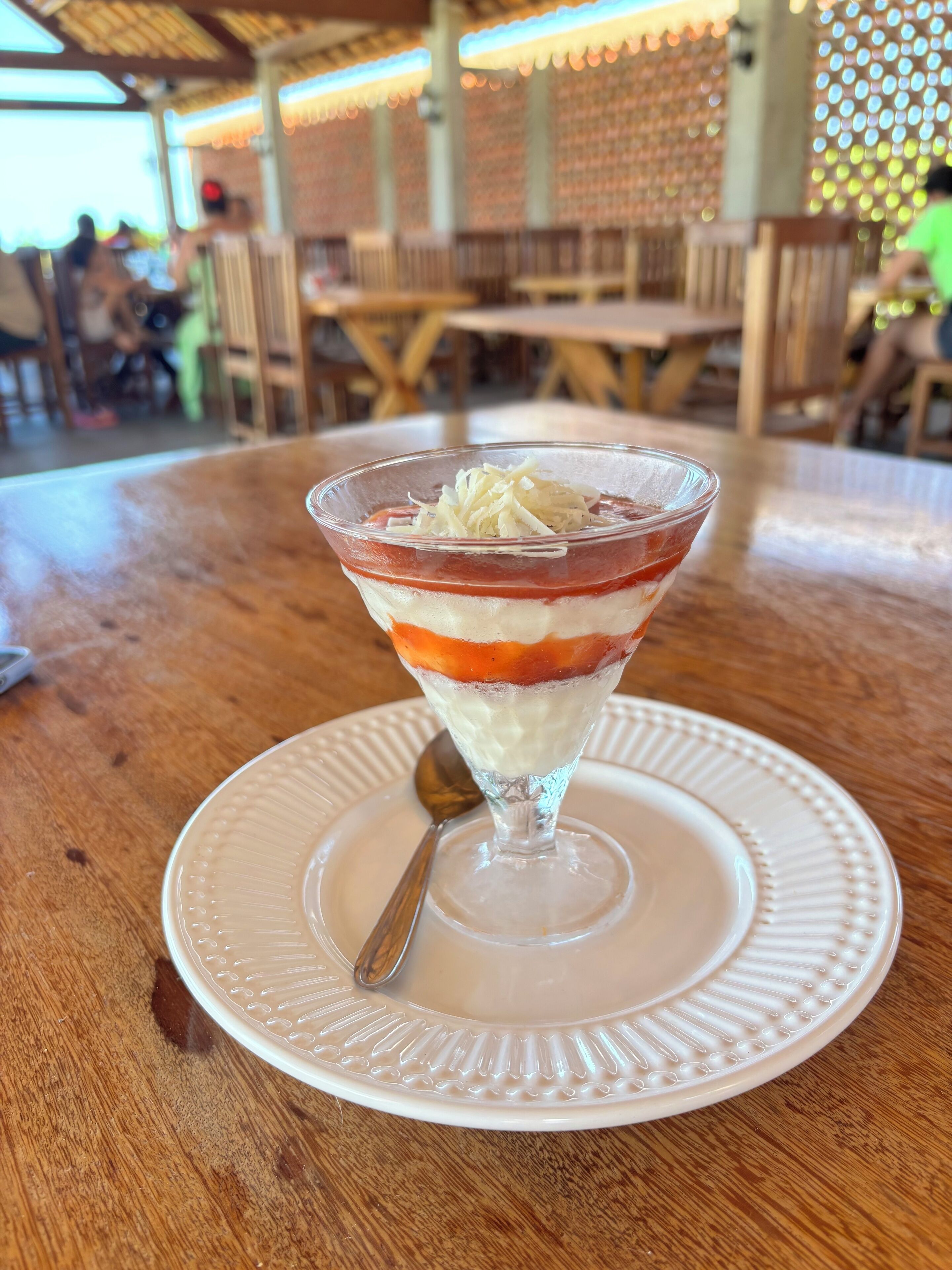 A dessert in a clear glass on a white plate with a spoon, placed on a wooden table.