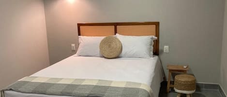 Una cama bien hecha con cabecera de madera, una mesita de noche y un taburete pequeño.