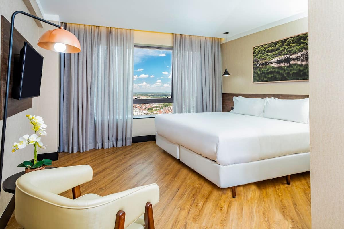 A hotel room with a bed, a desk with a chair, a TV, and a large window with curtains.