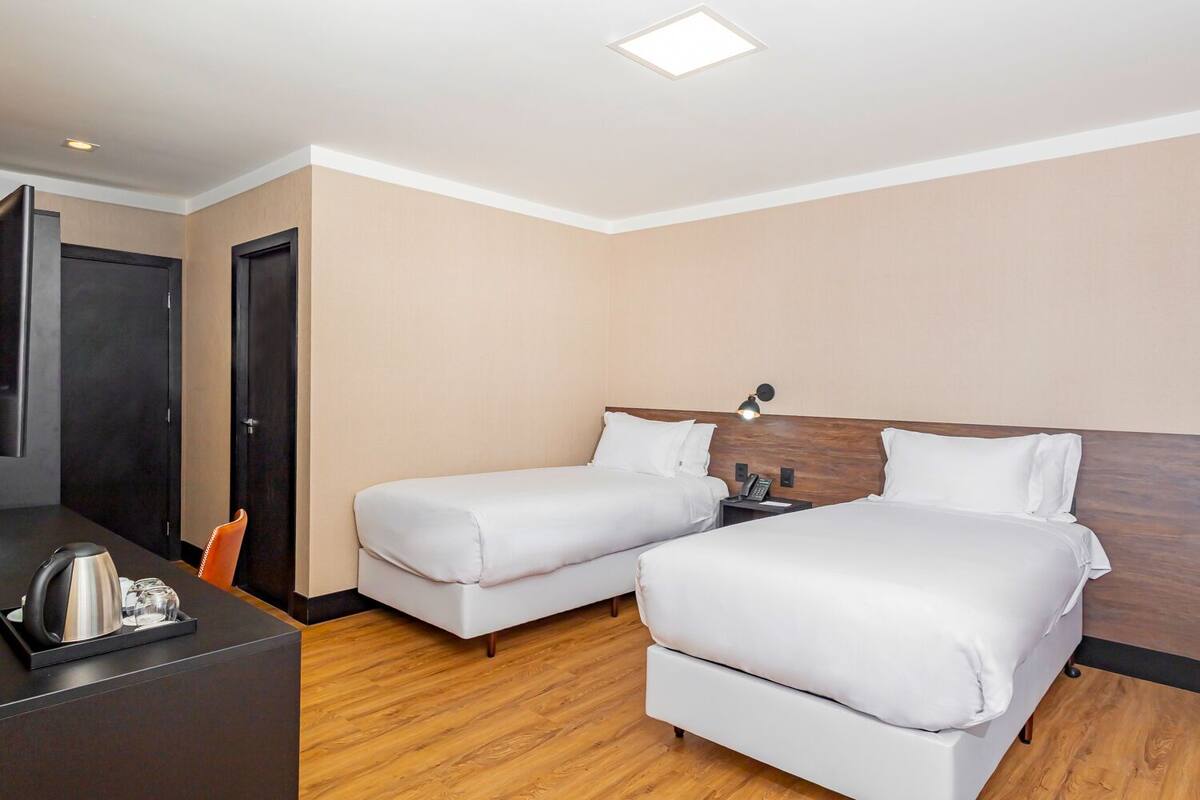 A hotel room with two beds, a desk with a kettle, a chair, and a door.