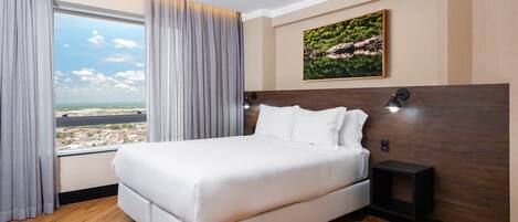 Ein Hotelzimmer mit einem großen Bett, Blick auf eine Stadtlandschaft, einem Bild an der Wand und einem Nachttisch mit Lampe.