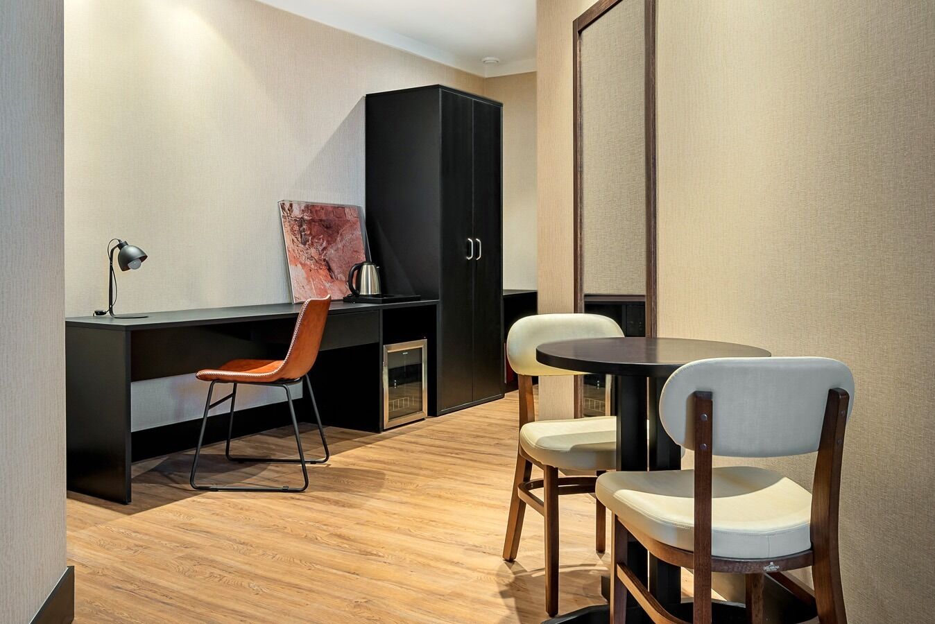 A modern hotel room with a wooden floor, a desk with a chair, a black wardrobe, and a round table with two chairs.