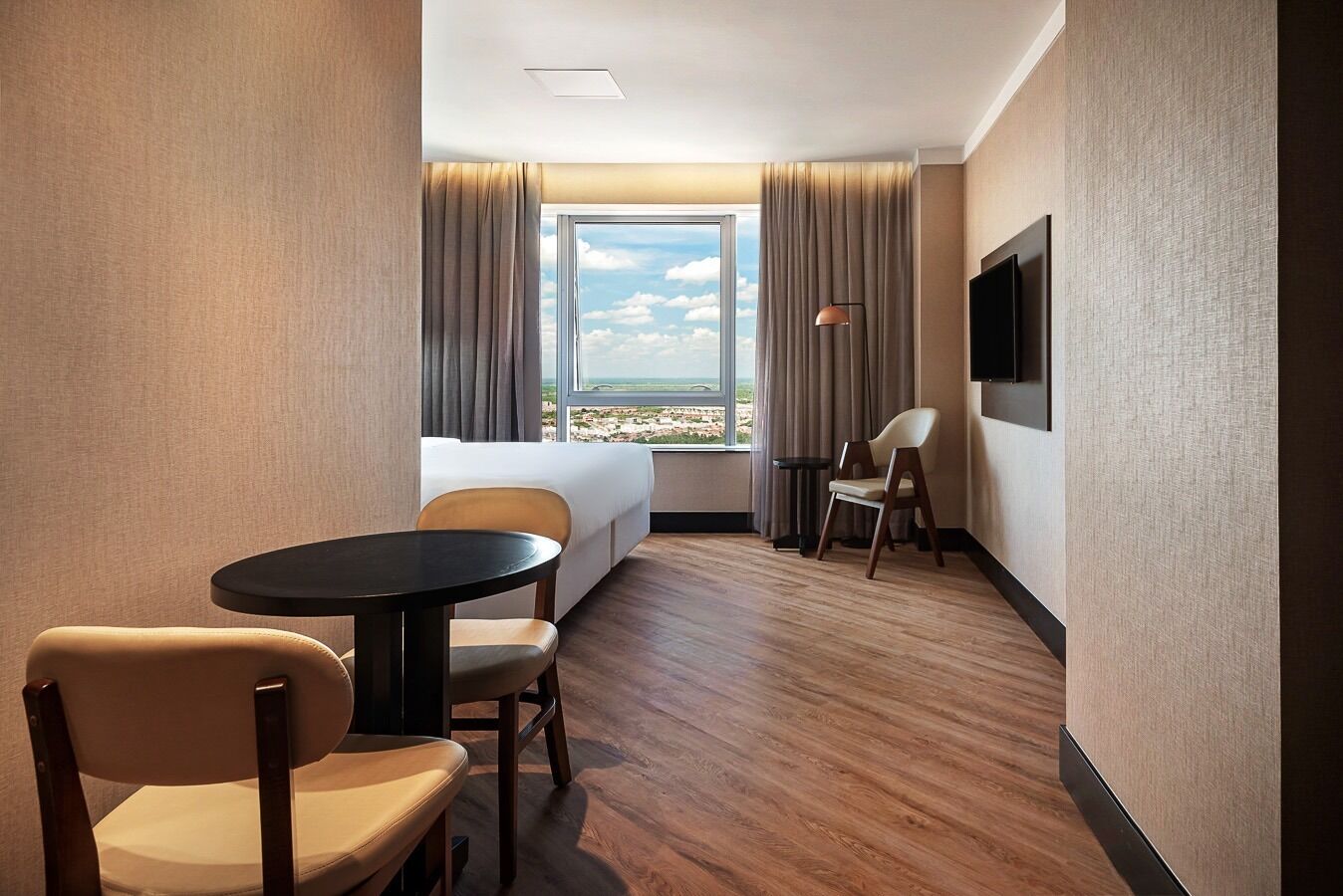 Habitación de hotel con cama, una mesa redonda, dos sillas, televisor y ventana con cortinas.