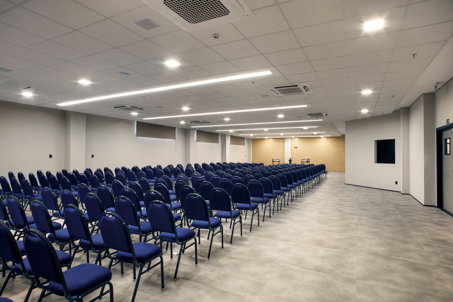 A conference room with rows of blue chairs, a wooden panel wall, and a light-colored floor.