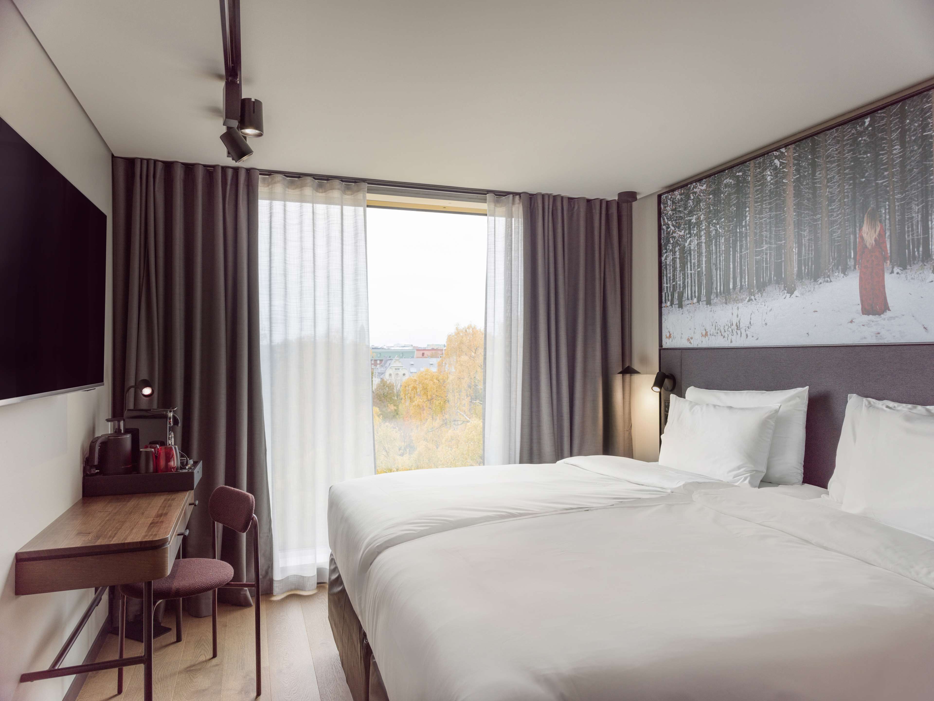 A hotel room with a bed, a desk, a chair, a TV, and a painting of a forest scene.