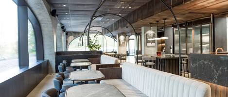 Un interior de restaurante moderno con un largo banco acolchado blanco, varias mesas redondas y una zona de bar.