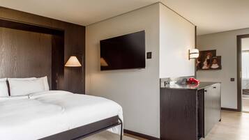 Quarto de hotel com cama, televisão na parede, escrivaninha com luminária e uma pequena cozinha equipada.