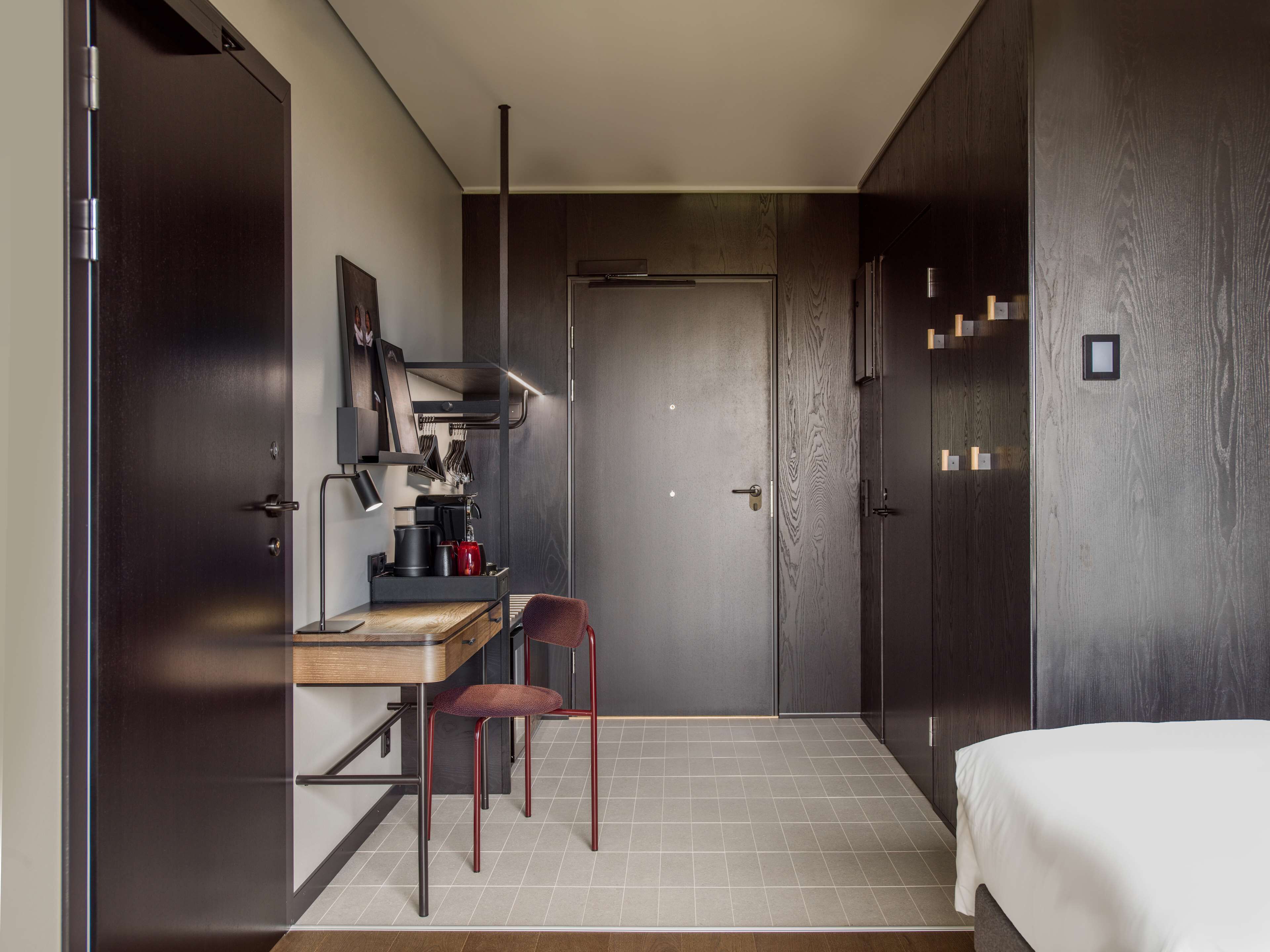 A compact hotel room with a bed, a desk with a chair, a kitchenette, and a wardrobe.
