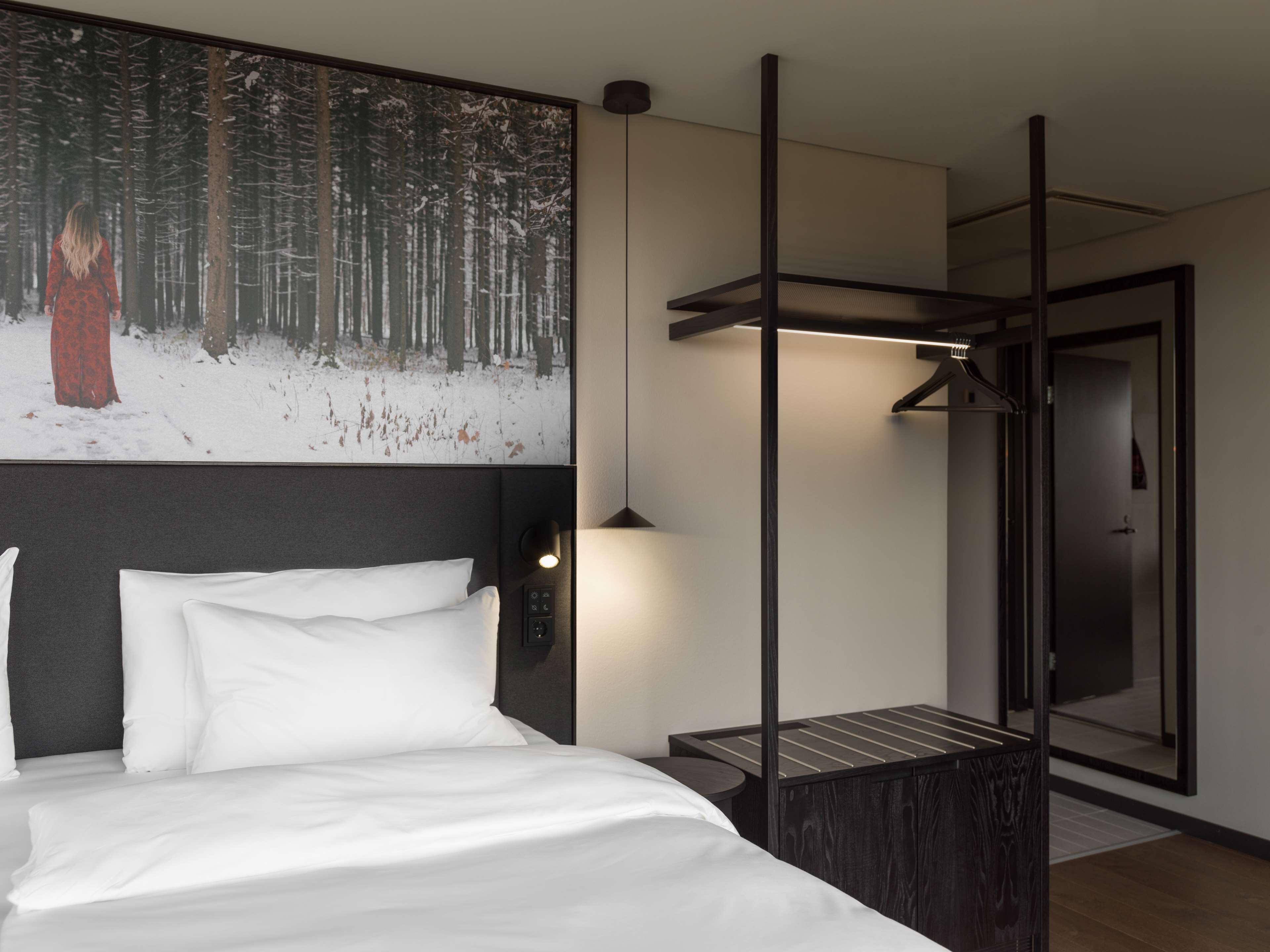 A modern hotel room with a large bed, a nightstand, a wall-mounted lamp, and a painting of a forest scene.