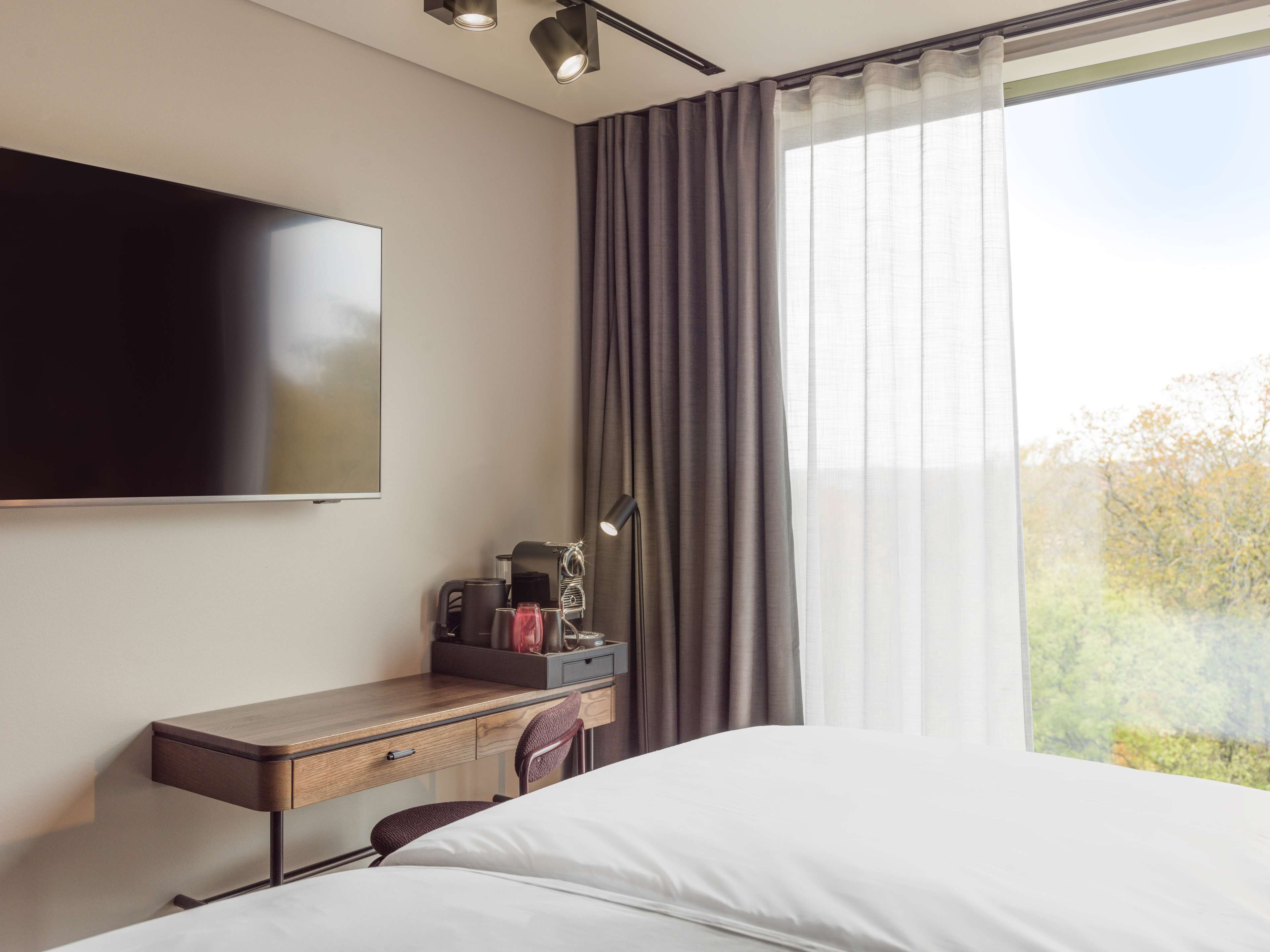 Habitación de hotel con cama, escritorio con silla, televisor de pantalla plana, cafetera y ventana con cortinas.