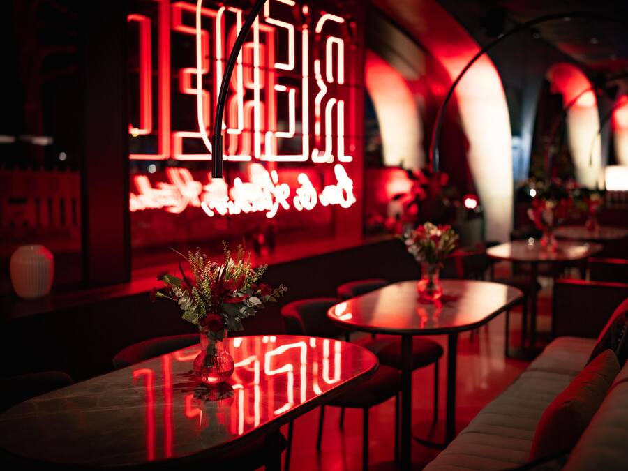 Un interior tenuemente iluminado con mesas, un sofá, un letrero de neón y un jarrón con flores.