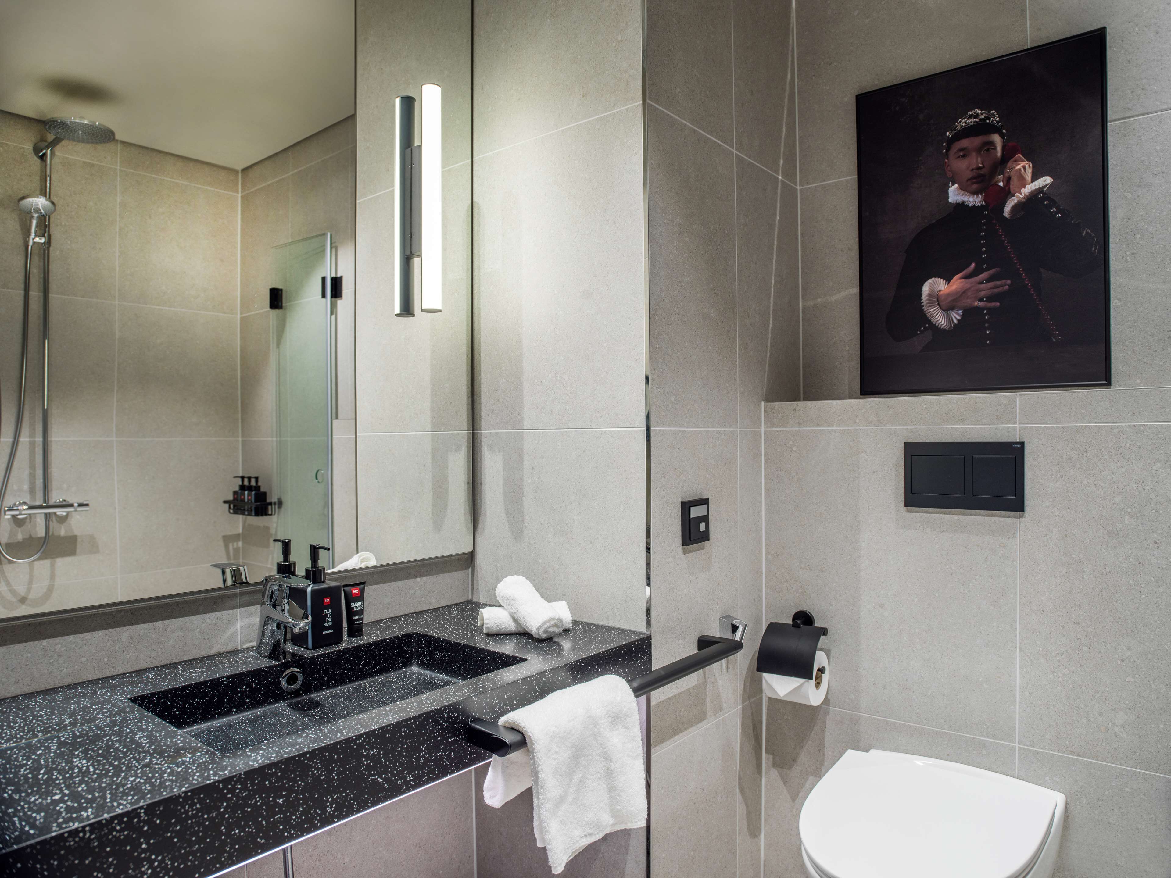 A modern bathroom with a large mirror, a shower, a sink with a black countertop, a toilet, and a framed portrait on the wall.