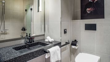 A modern bathroom with a large mirror, a shower, a sink with a black countertop, a toilet, and a framed portrait on the wall.