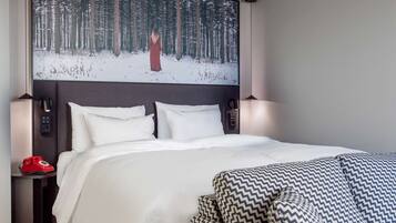 Una habitación de hotel moderna con una cama grande, un cabecero con una obra de arte de temática forestal, lámparas de noche, un teléfono giratorio rojo y un sillón con estampado.