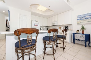 A modern kitchen with white cabinets, a central island, and bar stools with blue cushions. - 3BD Condo @ The Lighthouse 215 (Gulf Shores)
