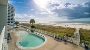 Zona de piscina con vista a la playa, una palmera y un edificio al fondo.