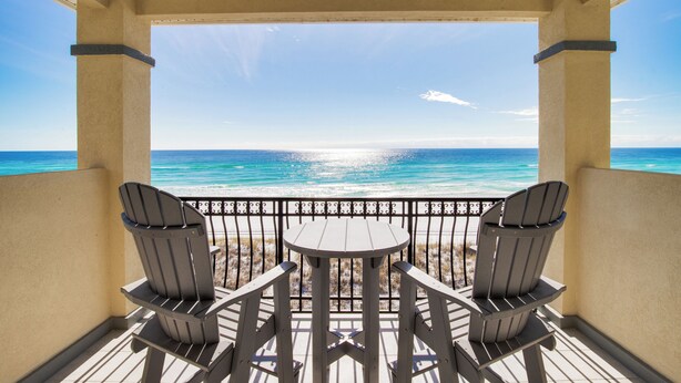 A balcony with two Adirondack chairs and a small table, overlooking a beach with clear blue water and a sunny sky. - Hot Tub - Destiny by the Sea Gated Community- 5-Star Reviews - On the Beach (Destin)