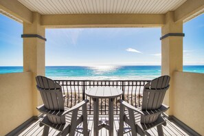 Een balkon met twee Adirondack-stoelen en een kleine tafel, met uitzicht op een strand met helderblauw water en een zonnige lucht.