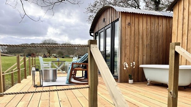 Una terraza de madera con barandilla de cuerda, una bañera blanca y una pequeña estructura de madera con puertas de cristal.