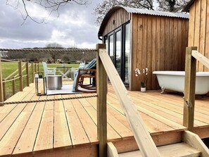 A wooden deck with a rope barrier, a white bathtub, and a small wooden structure with glass doors. - Boutique Shepherds Hut set in a wildflower meadow on a farm with Highland Cows (Pulborough)