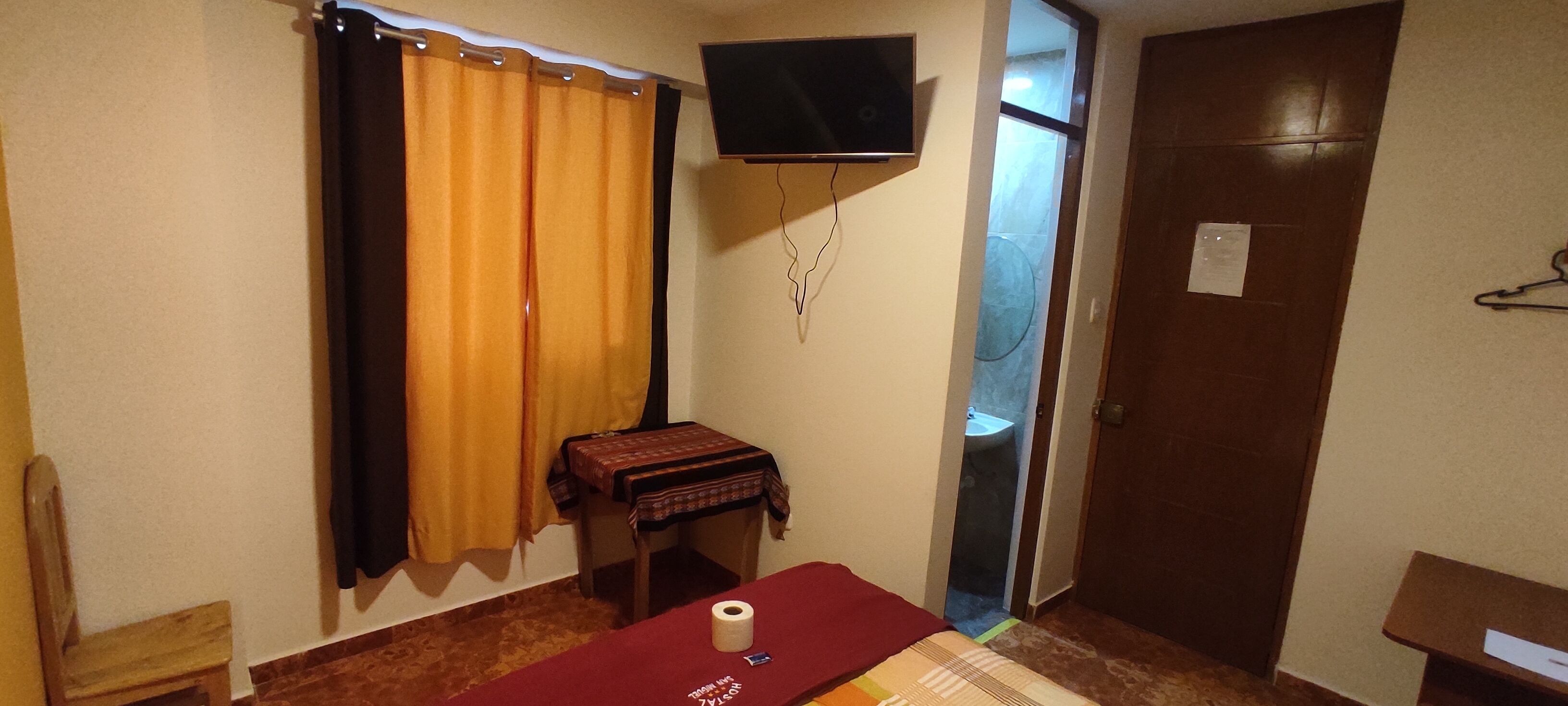 Ein Zimmer mit einem Bett, einem kleinen Tisch mit roter Tischdecke, einem an der Wand befestigten Fernseher, einer Tür und einem Fenster mit Vorhängen.