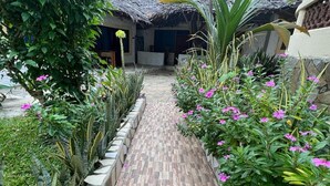 A pathway leading to a building with a thatched roof, surrounded by lush greenery and flowering plants.