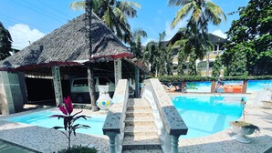 A swimming pool area with a thatched-roof structure, steps leading to the pool, and a mural in the background.