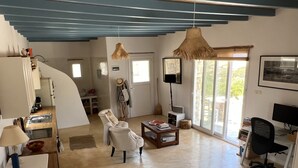 A spacious living area with a kitchen, a dining area, and a seating space with a sofa and armchairs. There are two hanging lights, a television, and a bookshelf. - Cosy open plan studio for couples or singles (Alyki)