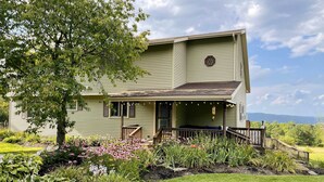 A two-story house with a porch, surrounded by a garden with various plants and flowers. - Spacious Hammondsport Home on 6 Acres w/ Lake View (Hammondsport)