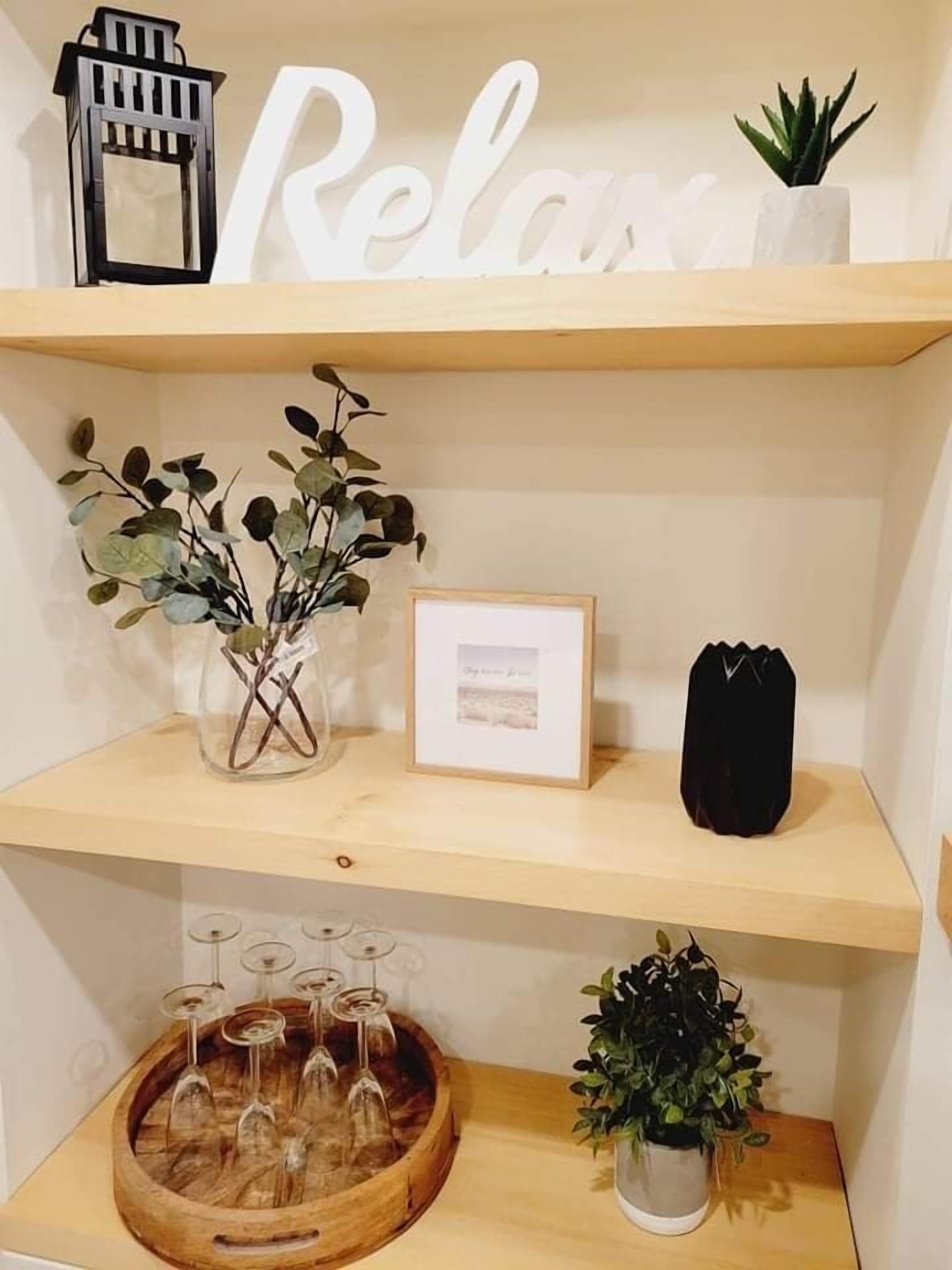 Une étagère avec une pancarte indiquant « Détendez-vous », un tableau encadré, une plante en pot et un plateau en bois contenant des verres à vin.