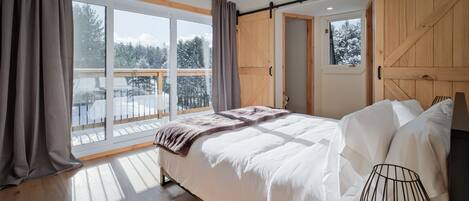 A bedroom with a large bed, a view of a snowy landscape through a sliding door, and a balcony with a wooden railing.