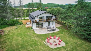 Une maison à deux étages avec un balcon, une terrasse dotée de meubles d’extérieur et un foyer entouré de chaises rouges.