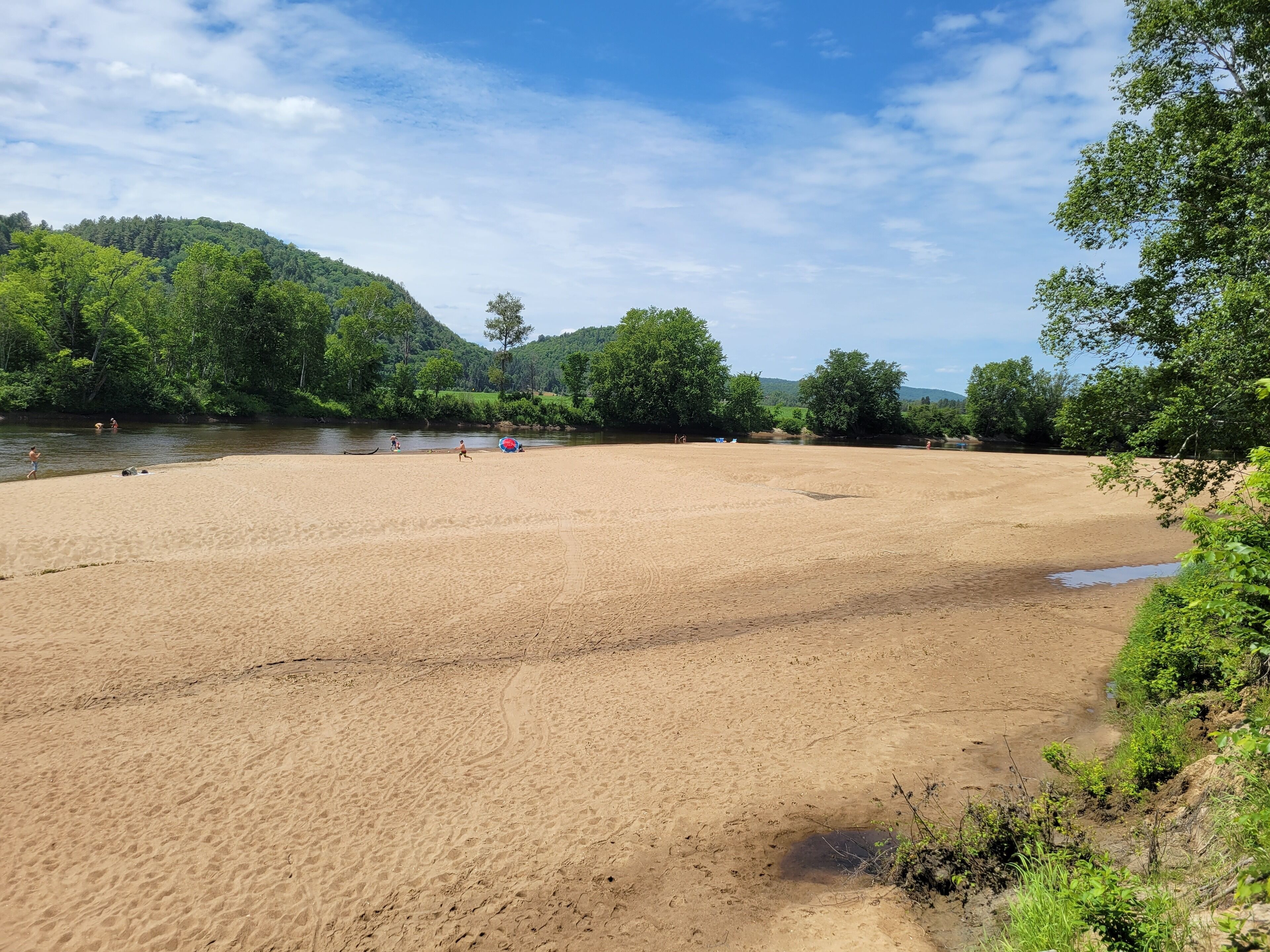 A sandy beach with a river and forested hills in the background.