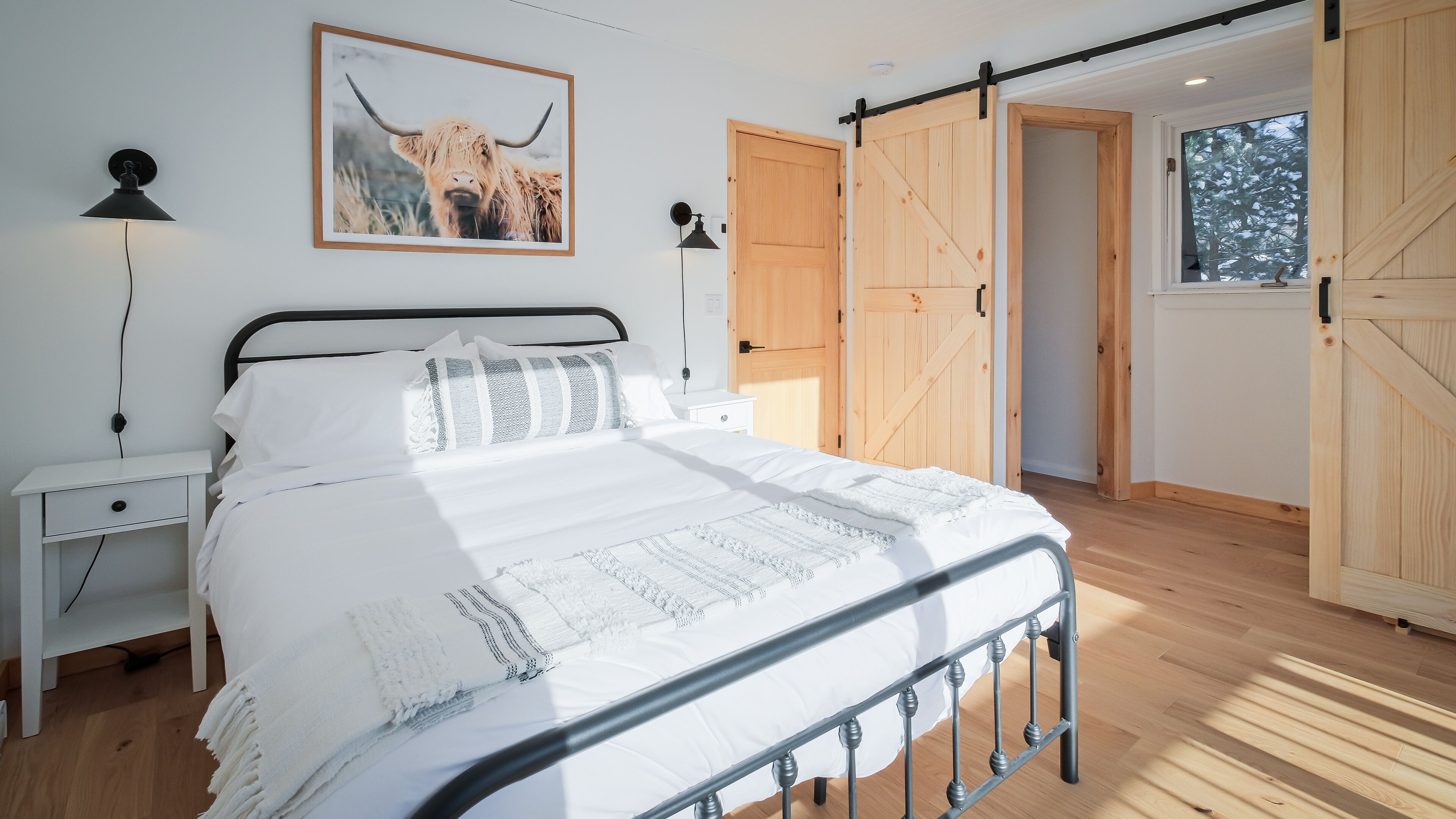 Une chambre à coucher avec un grand lit, une table de chevet, une lampe fixée au mur, une porte en bois et une grande fenêtre.