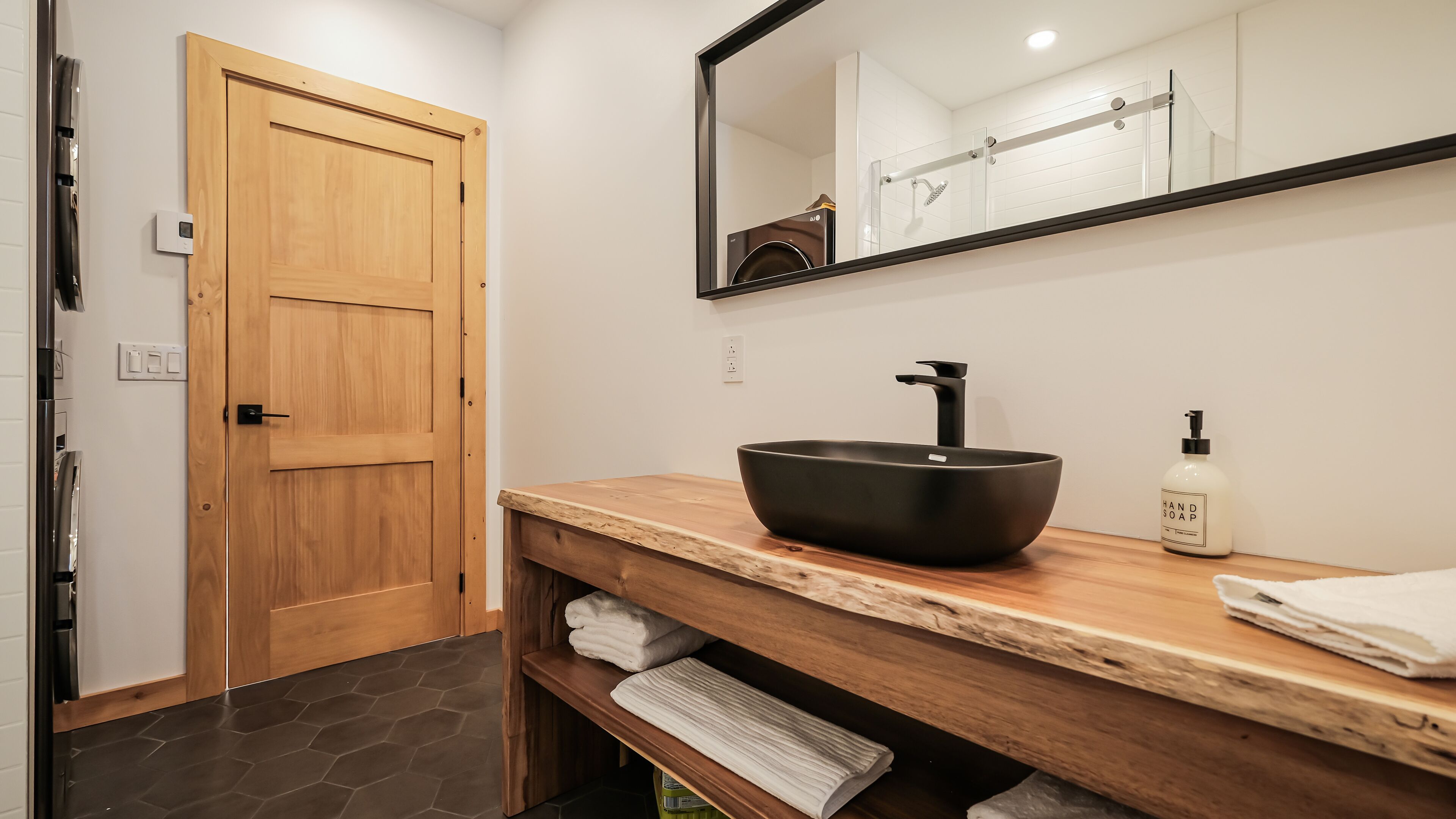 Une salle de bain avec une porte en bois, un lavabo noir, un miroir et un porte-serviettes.