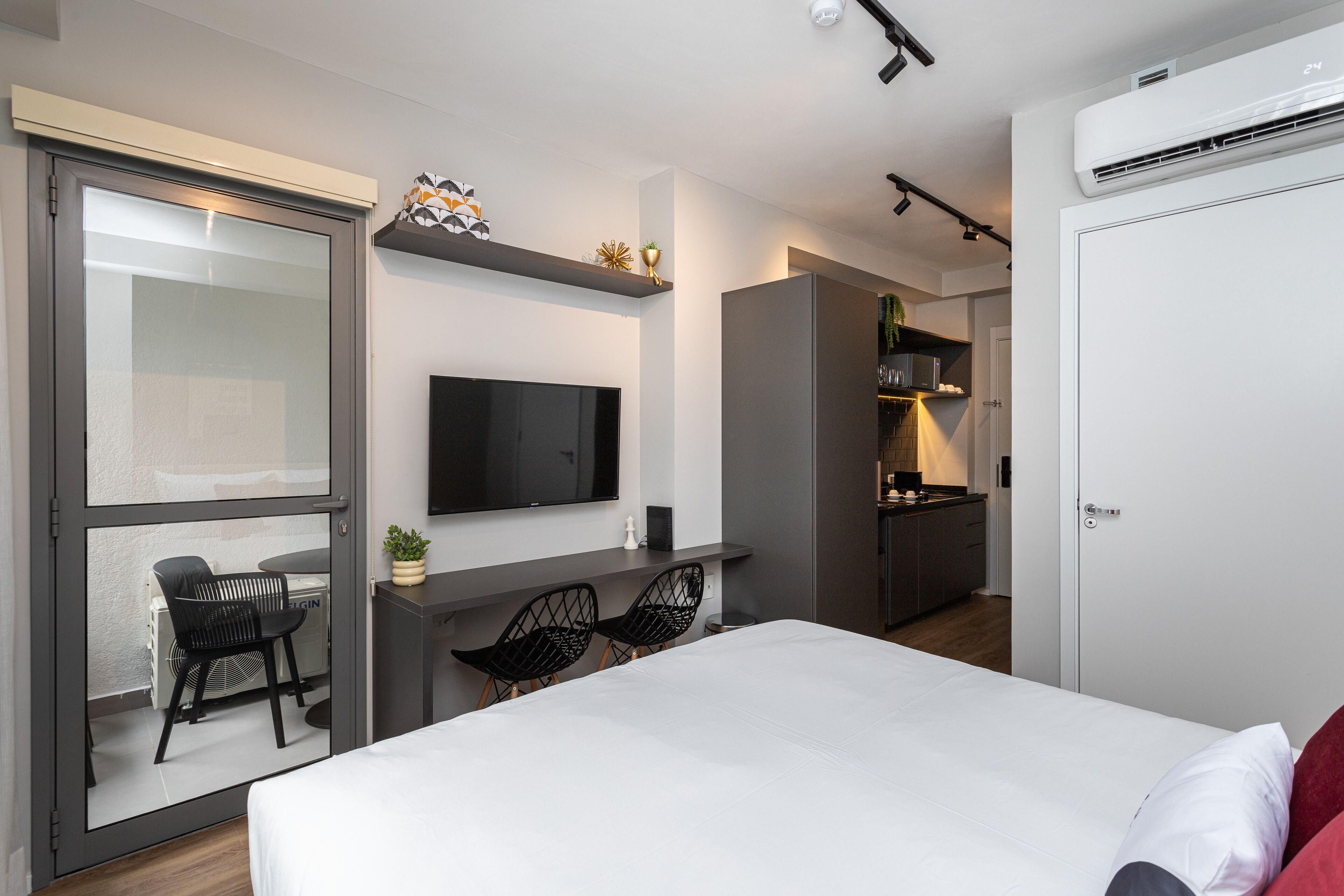 A modern hotel room with a large bed, a TV mounted on the wall, a small kitchen area, and a sliding door leading to an outdoor seating area.