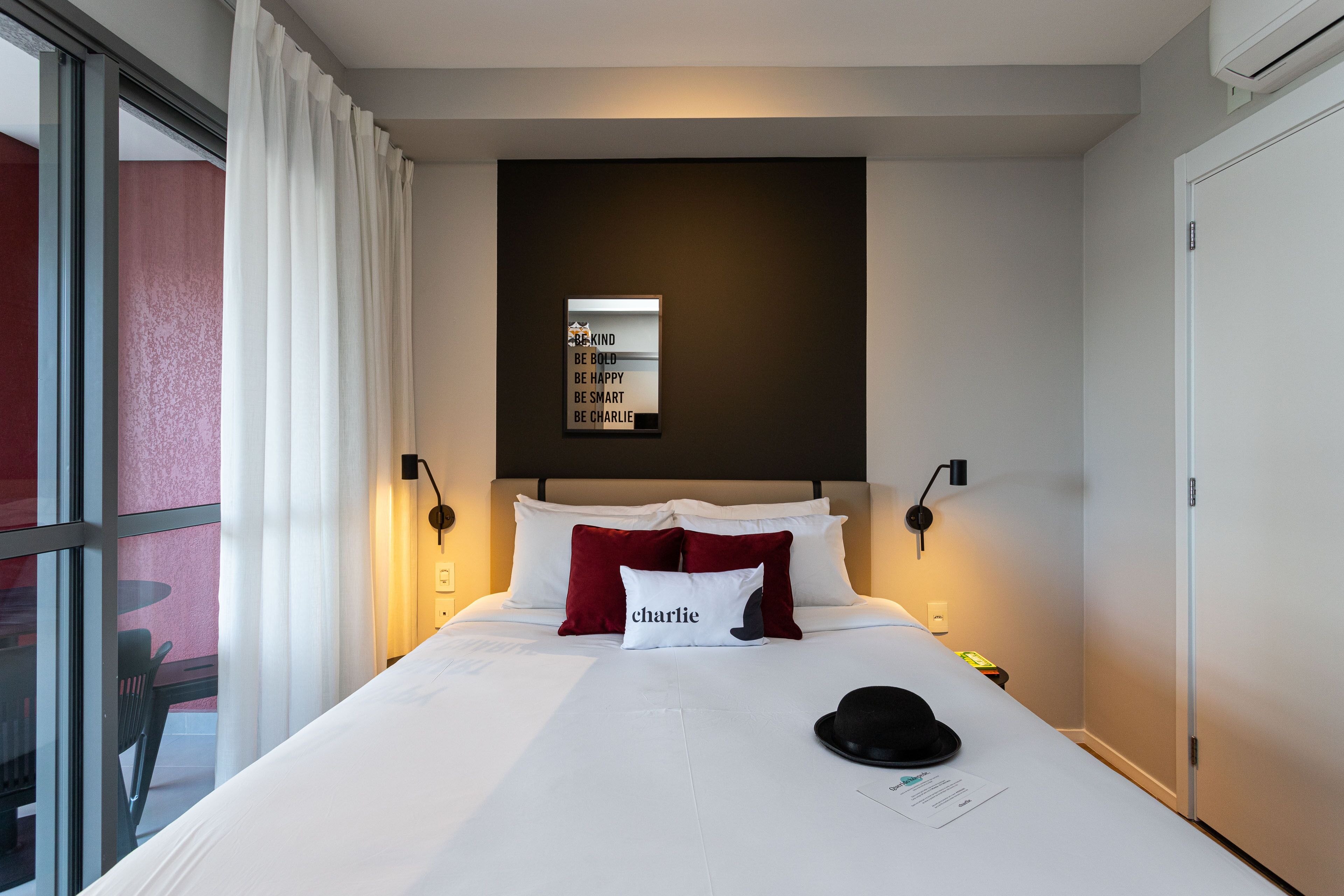 A hotel room with a bed, a black hat on the bed, a wall-mounted shelf with motivational quotes, and a window with curtains.