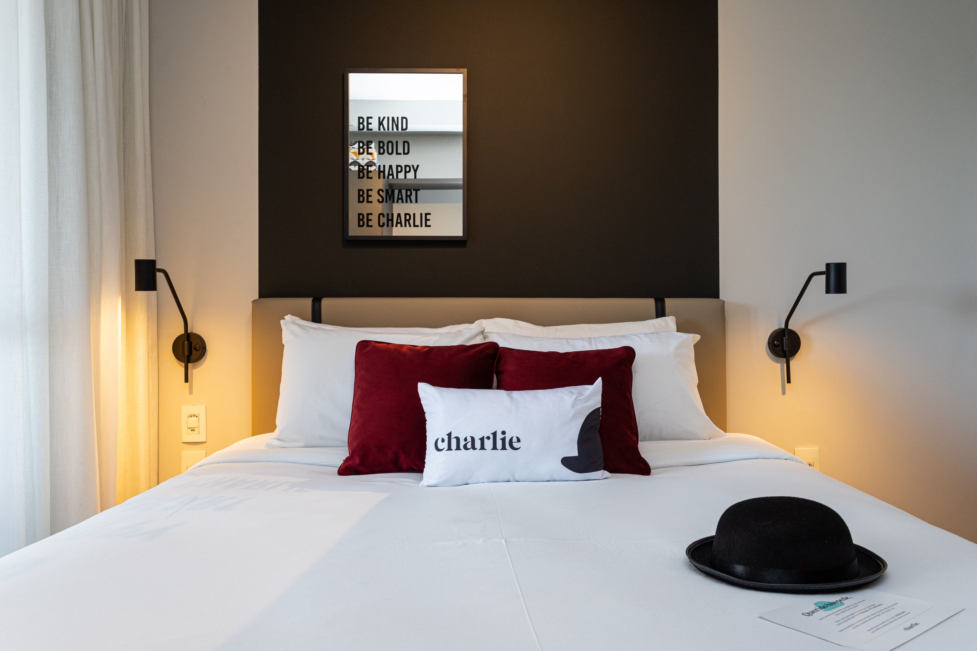 Ein Hotelzimmer mit einem Bett, einem Kopfkissen mit dem Namen "Charlie", einem an der Wand befestigten Schild mit motivierenden Sprüchen und einem schwarzen Hut auf dem Bett.
